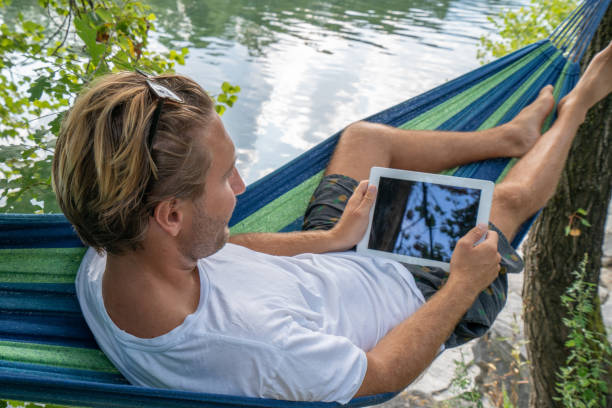 istock Young man using digital tablet on hammock, lying between trees by the river un Summer- People travel vacations technology on the go concept 1076374076