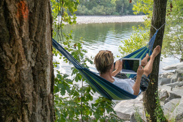 istock Young man using digital tablet on hammock, lying between trees by the river un Summer- People travel vacations technology on the go concept 1076375948