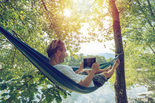 istock Young man using digital tablet on hammock, lying between trees by the river un Summer- People travel vacations technology on the go concept 1076376286