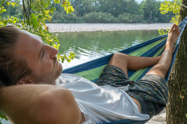 istock Young man relaxing on hammock by the river, hands behind head enjoying serene green environment. People travel relax concept in vacations 1076383332