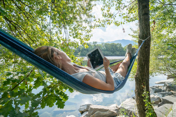川沿いの木の横になっているハンモックにデジタル タブレットを使用して若い女性国連夏-人々 の旅行休暇行くコンセプトの技術 - summer women hammock nature ストックフォトと画像