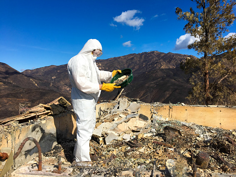 Man in hazmat suit sifting through ash and toxic waste after Woolsey wildfire. Burned Malibu (CA, USA) mountains in background, 2018.