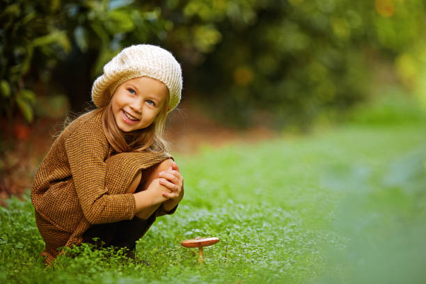 Side view of charming little girl in beret sitting on green lawn with growing mushroom smiling away