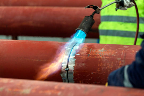 An Oilfield Worker Welds an End Cap to a Pipe as Sparks Fly Next to a Derrick at an Oil and Gas Drilling Pad Site on a Sunny Day
