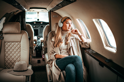 Young fashionable woman sitting in a private jet and listening to music through the headphones. She is looking through the window.