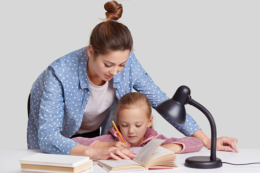 Horizontal shot of experienced young mother leans near her small child, helps to do home assignment, shows what to rewrite in book, surrounded with reading lamp, isolated over white background