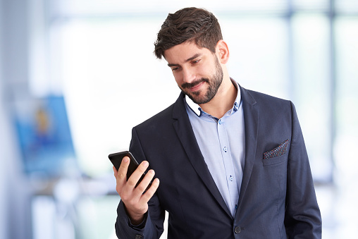 Smiling young businessman using his mobile phone and text messaging while standing in the office.