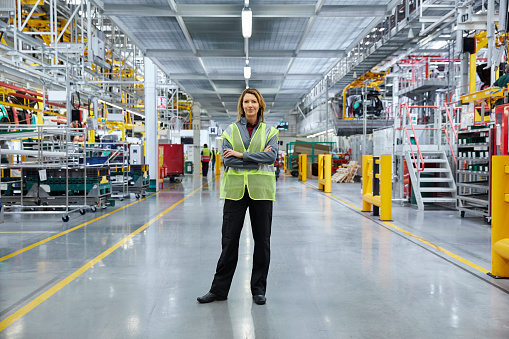 Portrait of mature female supervisor standing with arms crossed at automobile industry. Full length of confident manager is working at factory. She is wearing reflective clothing.