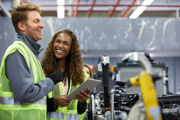 Smiling engineers standing with digital tablet Happy mechanical engineers holding digital tablet by car chassis at factory. Smiling colleagues are standing by car part at automobile industry. They are wearing reflective clothing. car engineer stock pictures, royalty-free photos & images