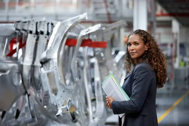 Portrait of confident manager standing by car chassis. Side view of young professional is holding documents. Engineer is working in automobile industry.