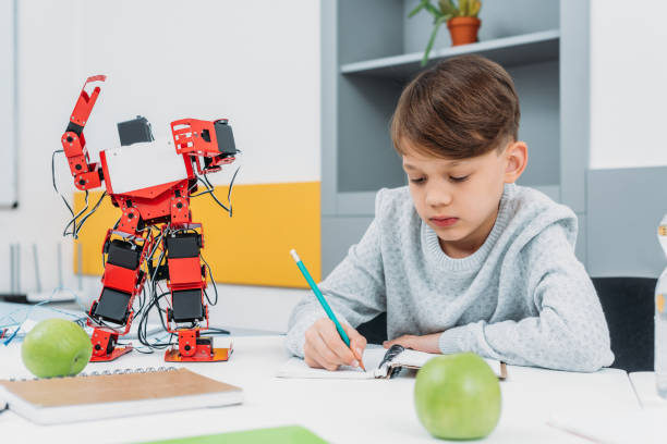 istock focused schoolboy sitting at desk with robot model and writing in notebook during STEM lesson 1076614122