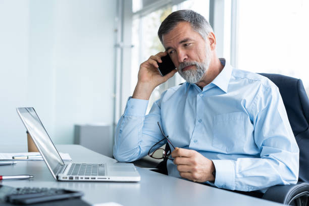 empresario senior ejecutivo utilizando su teléfono móvil y hablando wih alguien mientras trabajo laptop en la oficina. - reflection businessman business telephone fotografías e imágenes de stock