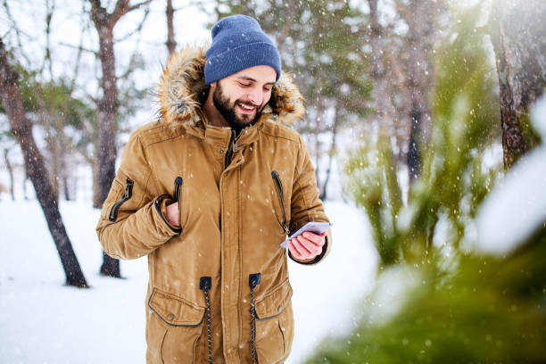 istock Smiling bearded man wears warm winter clothes and using smartphone with fast internet connection in country side. Handsome man texting with cellphone and using apps in forest. Snowfall in woods. 1076631752