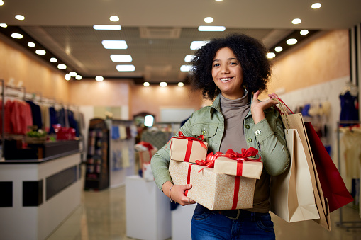 Happy african american girl shopping gifts in mall on christmas sale. New year holidays shopping idea concept. Smiling mixed race woman with colorful paper gift boxes wearing christmas hat.