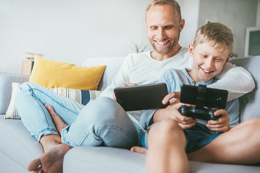 Father and son game players funs sit together at home on cozy sofa, playing the games using the tablet and gamepad. Polurar gadgets using concept
