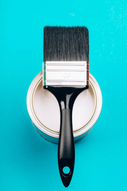 Brush with black handle on open can of white paint on blue background.