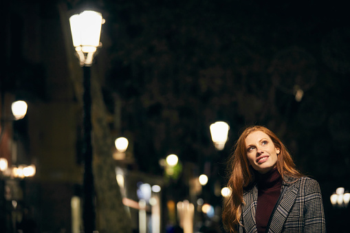 Thoughtful smiling woman looking away. Young female is standing by illuminated street light. She is in city at night.