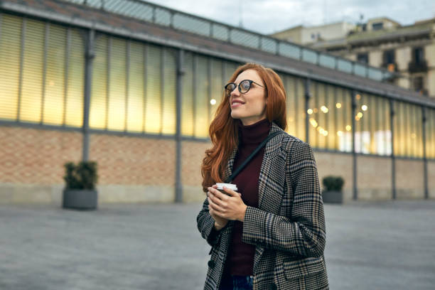 Thoughtful smiling woman holding disposable cup Smiling woman holding coffee cup. Thoughtful female is looking away at city. She is in warm clothing. winter fashion stock pictures, royalty-free photos & images