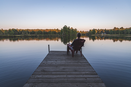 The image displays a young man relaxing on an Adirondack chair and looking at a calm river at sunset. The image was taken in Muskoka, Ontario, Canada.