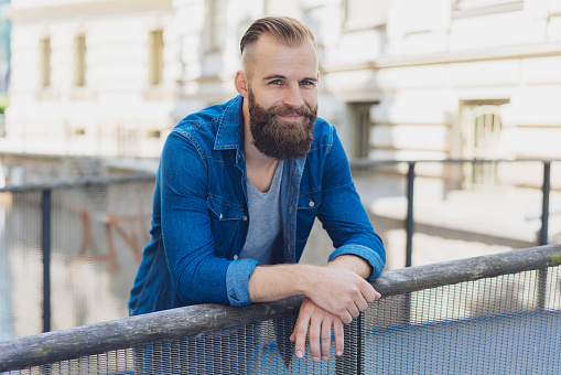 Smiling friendly bearded young man standing on a bridge leaning on the railing over a canal or river