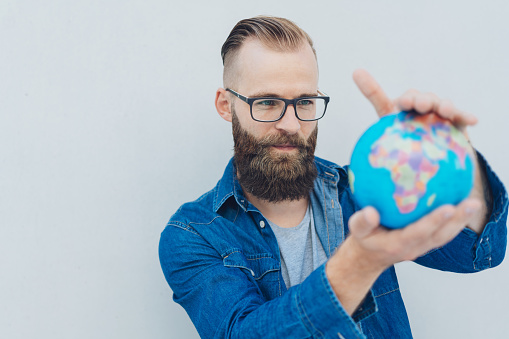 Bearded man wearing glasses holding up a world globe studying it with a look of concentration in a conceptual image of travel or conservation