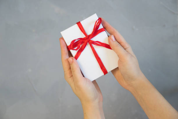istock Woman hands holding present box with red bow on pastel pink background with multicolored confetti. Flat lay style. 1076707318