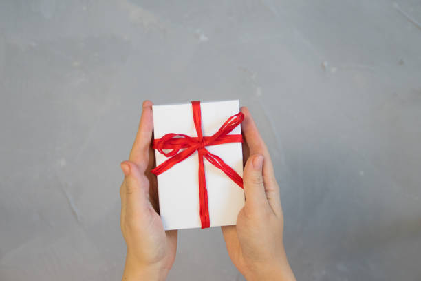 istock Woman hands holding present box with red bow on pastel pink background with multicolored confetti. Flat lay style. 1076707346