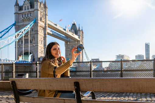 Beautiful female London traveler takes a selfie picture with her phone in front of the Tower Bridge on a sunny day