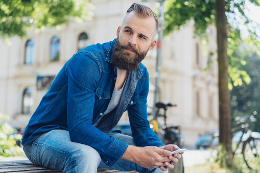 Street portrait of young bearded man wearing denim shirt sitting on bench with mobile phone