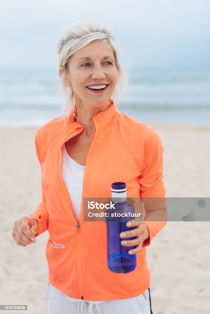 Blond woman exercising on a beach carrying water Blond woman exercising on a beach carrying a blue bottle of water looking to the side with a beaming happy smile 60-69 Years Stock Photo