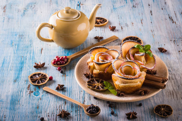 vertical photo with crispy dessert. sweet is made from apple slices and puff pastry. dessert is called apple roses. several herbs and spices are spilled around with powder sugar. - powder puff imagens e fotografias de stock