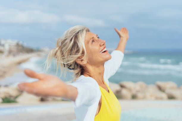 Joyful woman enjoying the freedom of the beach Joyful woman enjoying the freedom of the beach standing with open arms and a happy smile looking up towards the sky senior women stock pictures, royalty-free photos & images