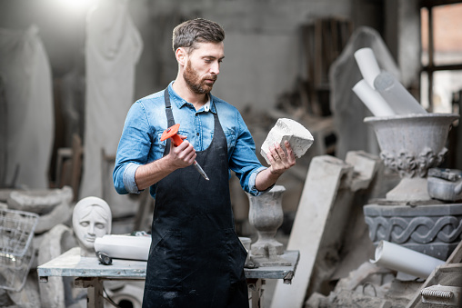 Portrait of a handsome sculptor in blue t-shirt and apron holding a piece of stone in the studio