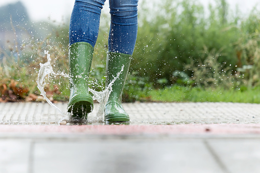 Close-up of woman in green rubber boots jumping on the puddle water in the street.