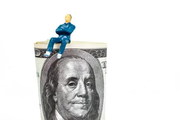 The concept of a successful businessman or stockbroker. A toy man is sitting on a hundred dollar bill against a white background.