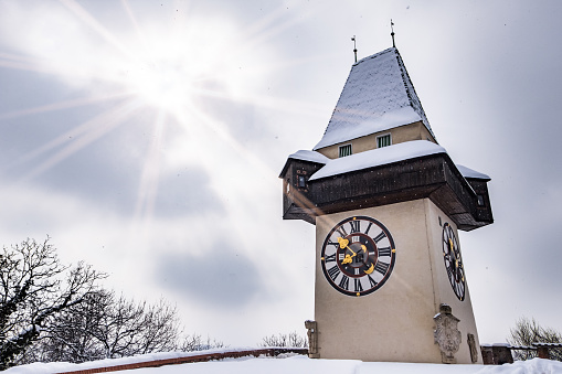 Sun flares over snow covered Uhrturm clocktower landmark of city Graz on hill Schlossberg in winter