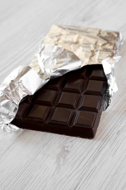 istock Dark chocolate in a wrapper over white wooden background, side view. 1076857946