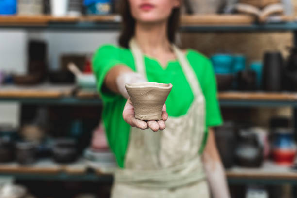 Master class concept. Blurred lady in her casual green t-shirt workwear she stand inside workspace show on camera small ceramics cup in hand focus on front object