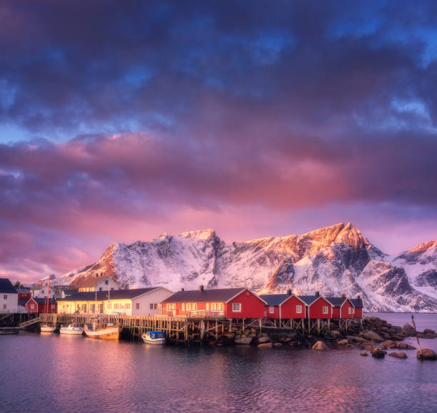 美しい漁村、日の出、ロフォーテン諸島ノルウェー船。住宅、雪山、海、船、カラフルな空の雲と冬の風景。ノルウェーの伝統的な赤がりと岩 - snow winter coastline sunrise ストックフォトと画像