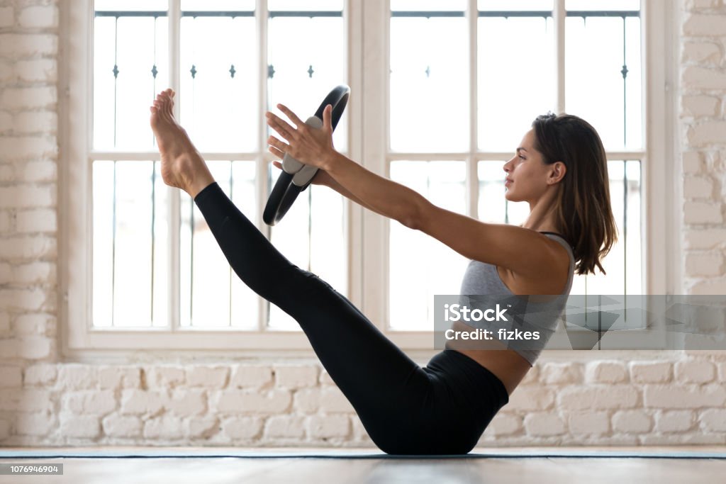 Young woman practicing yoga, Paripurna Navasana exercise with a ring Young sporty attractive woman doing toning pilates exercise for abs, balance Paripurna Navasana pose with pilates magic circle in hands, working out wearing sportswear, indoor full length, yoga studio Pilates Stock Photo