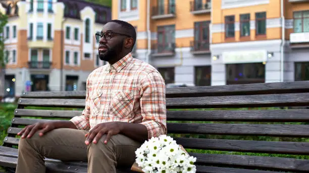 Young male waiting for woman, sitting on park bench with flowers, blind date