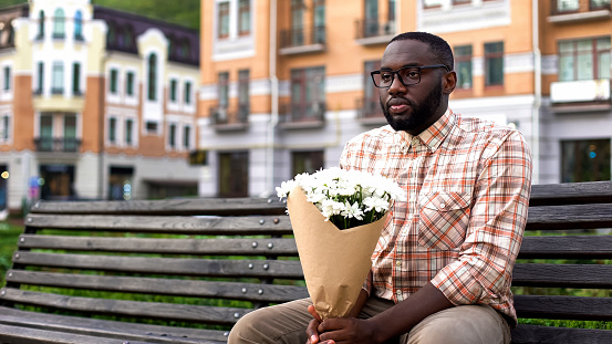 African man sitting lonely on city bench, holding flower bouquet, failed date