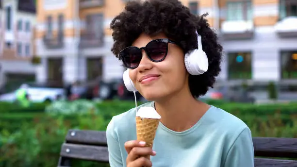 Inspired lady enjoying music in headphones, eating ice-cream on bench, leisure