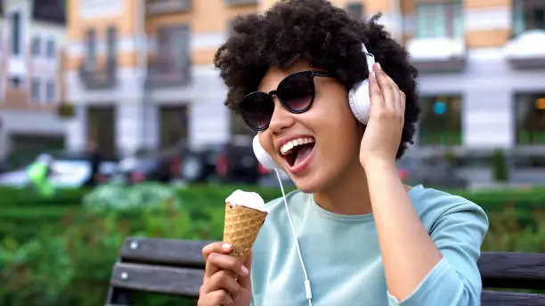 Afro-american woman enjoying music in headphones, eating ice-cream on bench