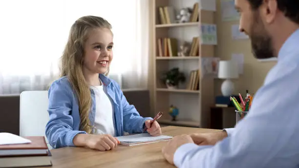 Father helping his daughter with homework, female kid sincerely smiling to dad