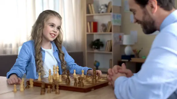 Cute girl smiling to father playing chess, first victory in game, family leisure