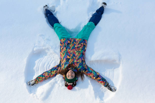 high angle view of happy girl lying on snow and moving her arms and legs up and down creating a snow angel figure. smiling woman lying on snow in winter holiday with copy space - 13427 imagens e fotografias de stock