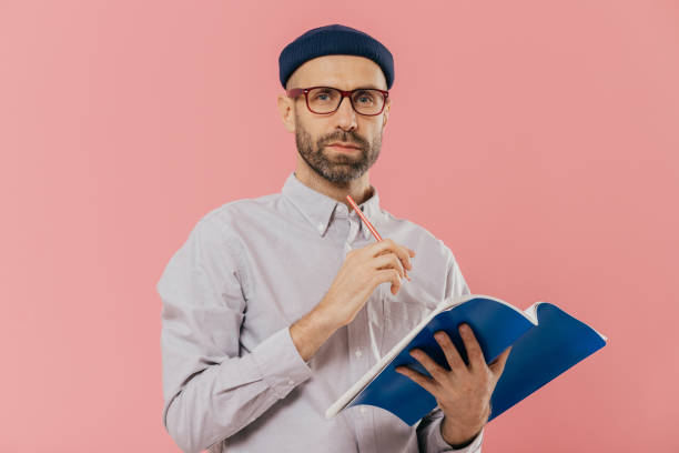 istock Isolated shot of attractive young male holds textbook, makes notes in organizer, dressed formally. Clever student prepares for examination or does home assignment, isolated over pink studio wall 1077061784