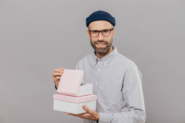 istock Handsome European man with thick stubble, pleasant smile, holds boxes in hands, opens gift with happiness, wears formal white shirt and headgear, isolated over grey background. Parcel in mans hands 1077061900