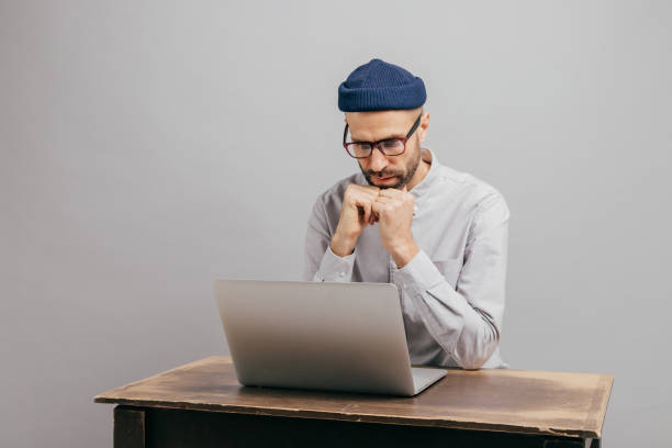 istock Serious male workaholic sits at desktop with modern electronic device, watches video, prepares financial report, concentrated in laptop computer, wears white shirt, optical spectacles, models indoor 1077061912
